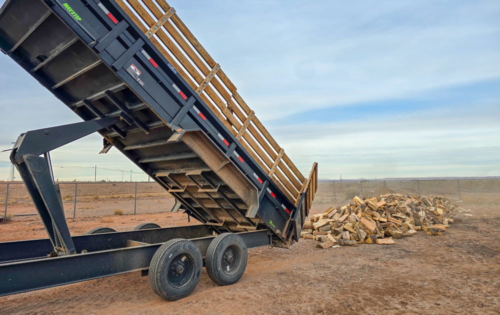 A pile of cut logs sits next to a large trailer. 