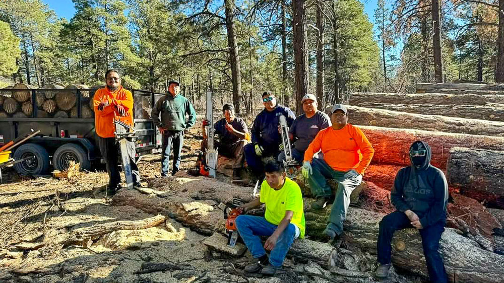 Several men sitting and standing next to a stack of timber. 