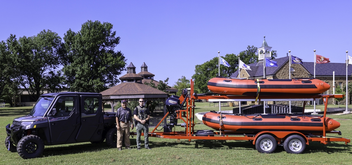 Two men stand in front of a utility terrain vehicle attached to a trailer that holds two pontoon rescue boats. 