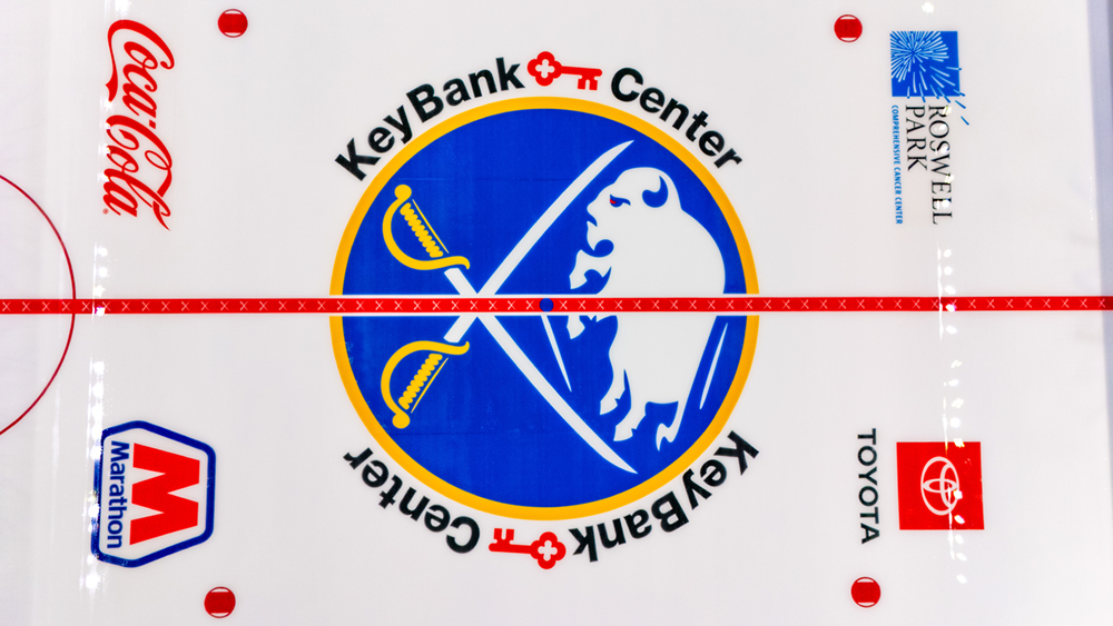 A large letter M and additional lettering with a buffalo logo at center ice on a hockey rink, crossed by a red center line.