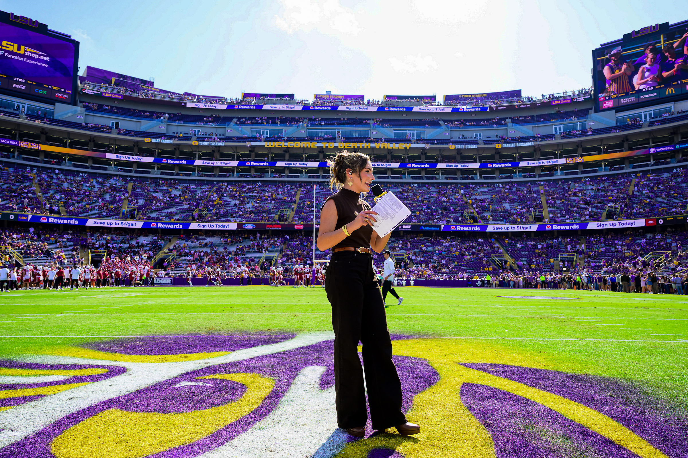 A female reporter stands on the turf inside a sports stadium filled with fans.