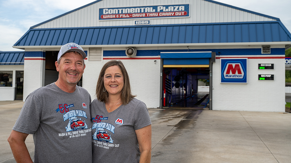 A man and woman stand outside a gas station and car wash for a photo.