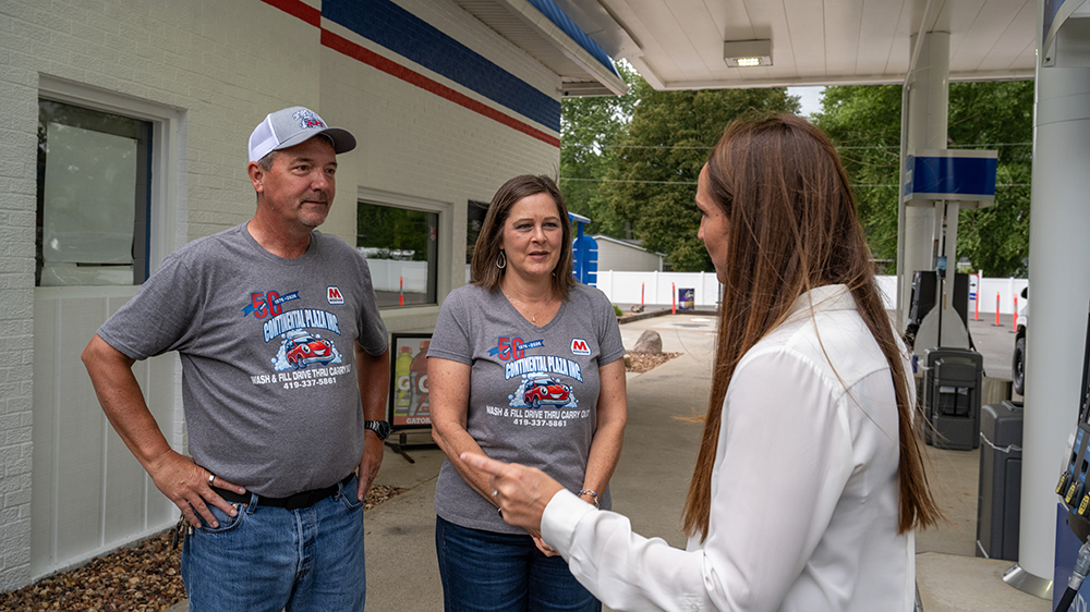 Three people have a conversation outside a gas station.