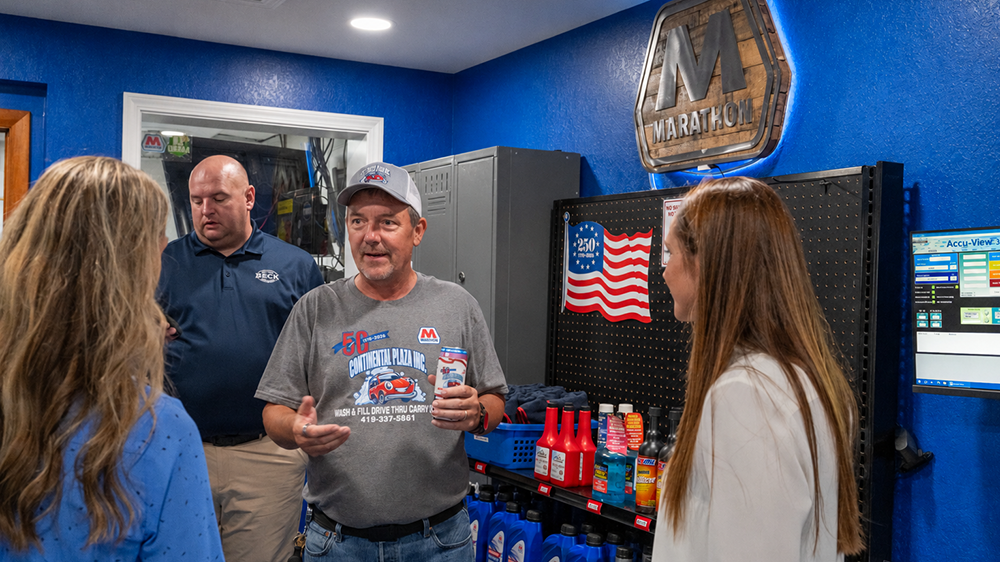 People talk inside a store with motor oil and an American flag in the background.