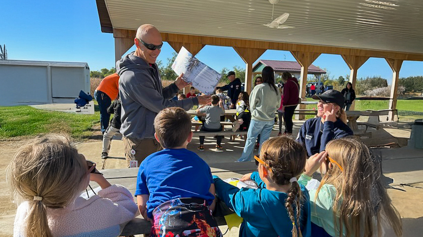 A man holds a book and reads to children outside under a pavilion at a nature park.