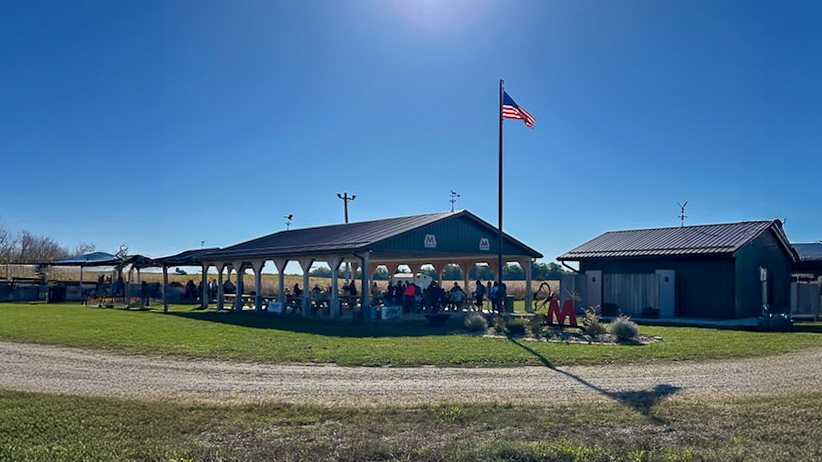 A park shelter on a sunny day with people standing and sitting underneath it, and an American flag flying next to the shelter.