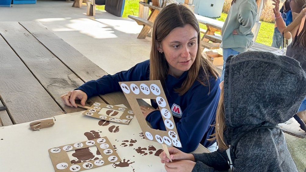 A woman and a child talk at a table with artwork and paint supplies under a pavilion at a nature park.