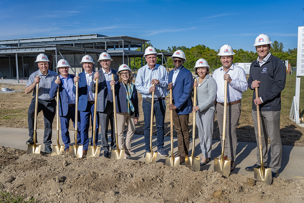 A group of people wearing hard hats use shovels during a groundbreaking ceremony.