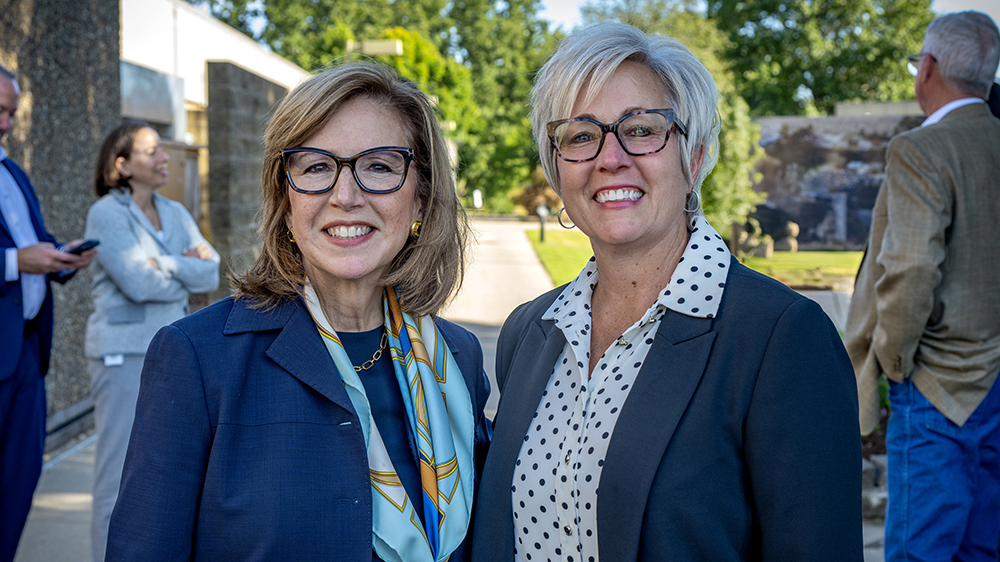 Two women stand together and smile for a photo outside a college building.