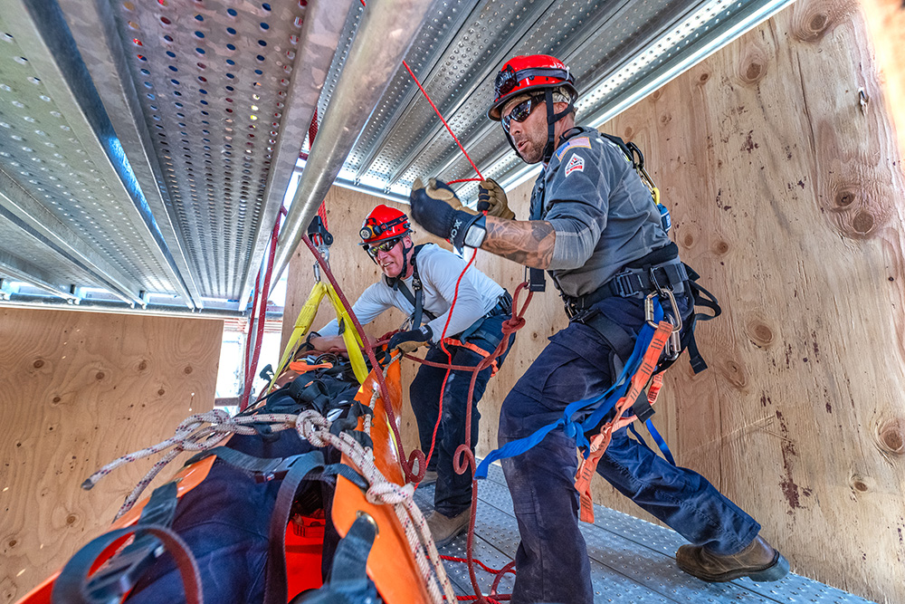 Two men on a platform use ropes to lower a man in a rescue basket.