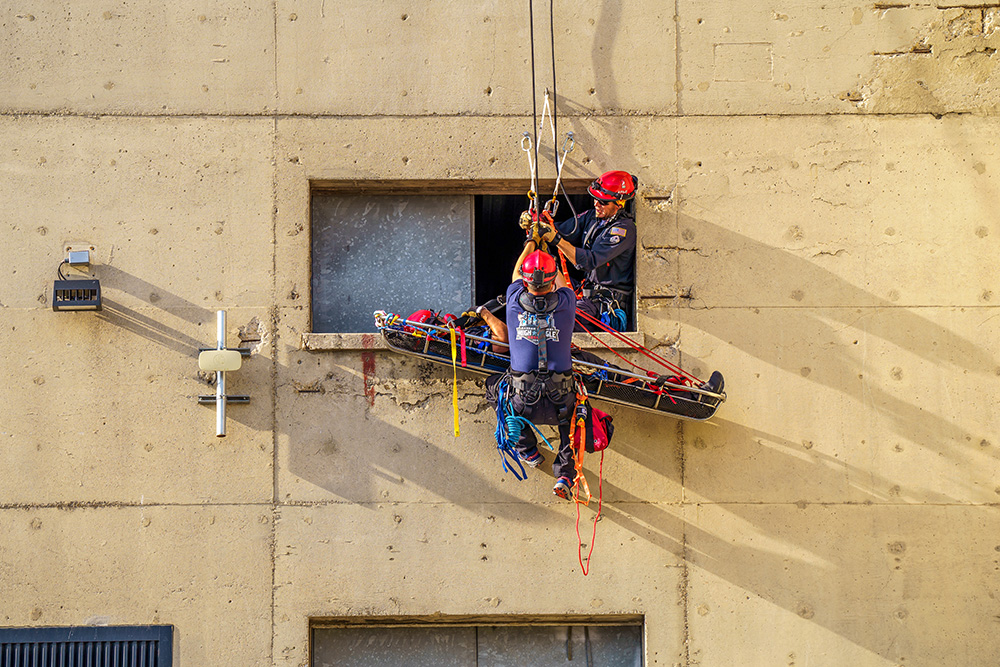 A man uses harnesses and ropes to lower another man in a rescue basket down the side of a building.