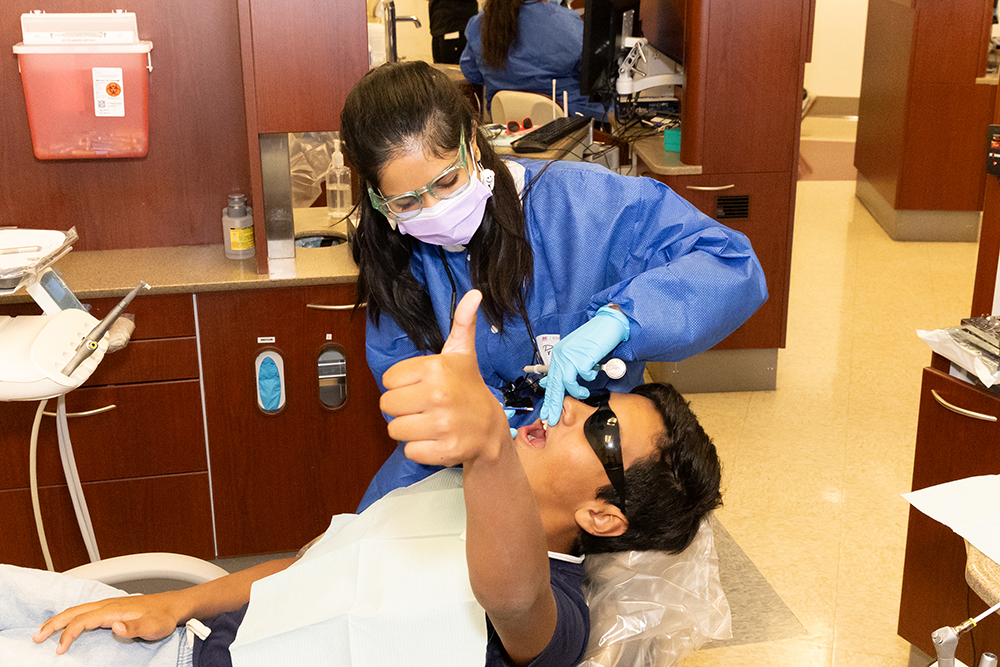 A teenage boy lies in a dental chair while a dental school student uses an instrument to complete a procedure. 