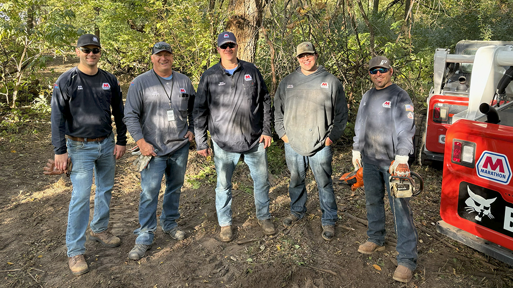 Five men stand together outdoors in the sun, smiling for a group photo.