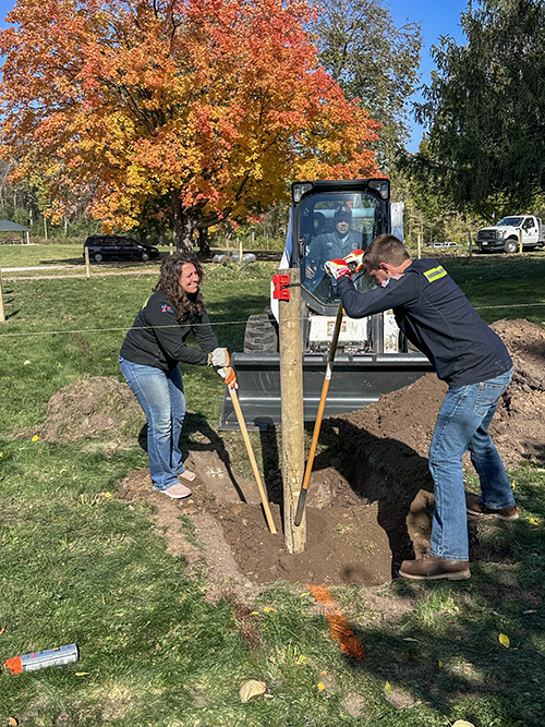: Two people outdoors installing a fence post, with one operating a skid loader.