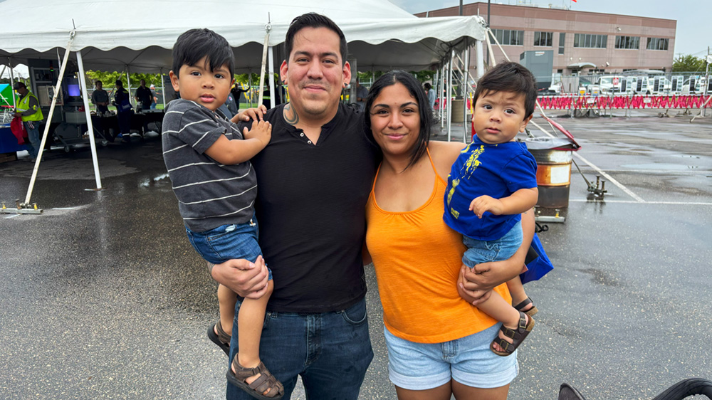 Parents holding two children stand at the fair with a tent in the background.