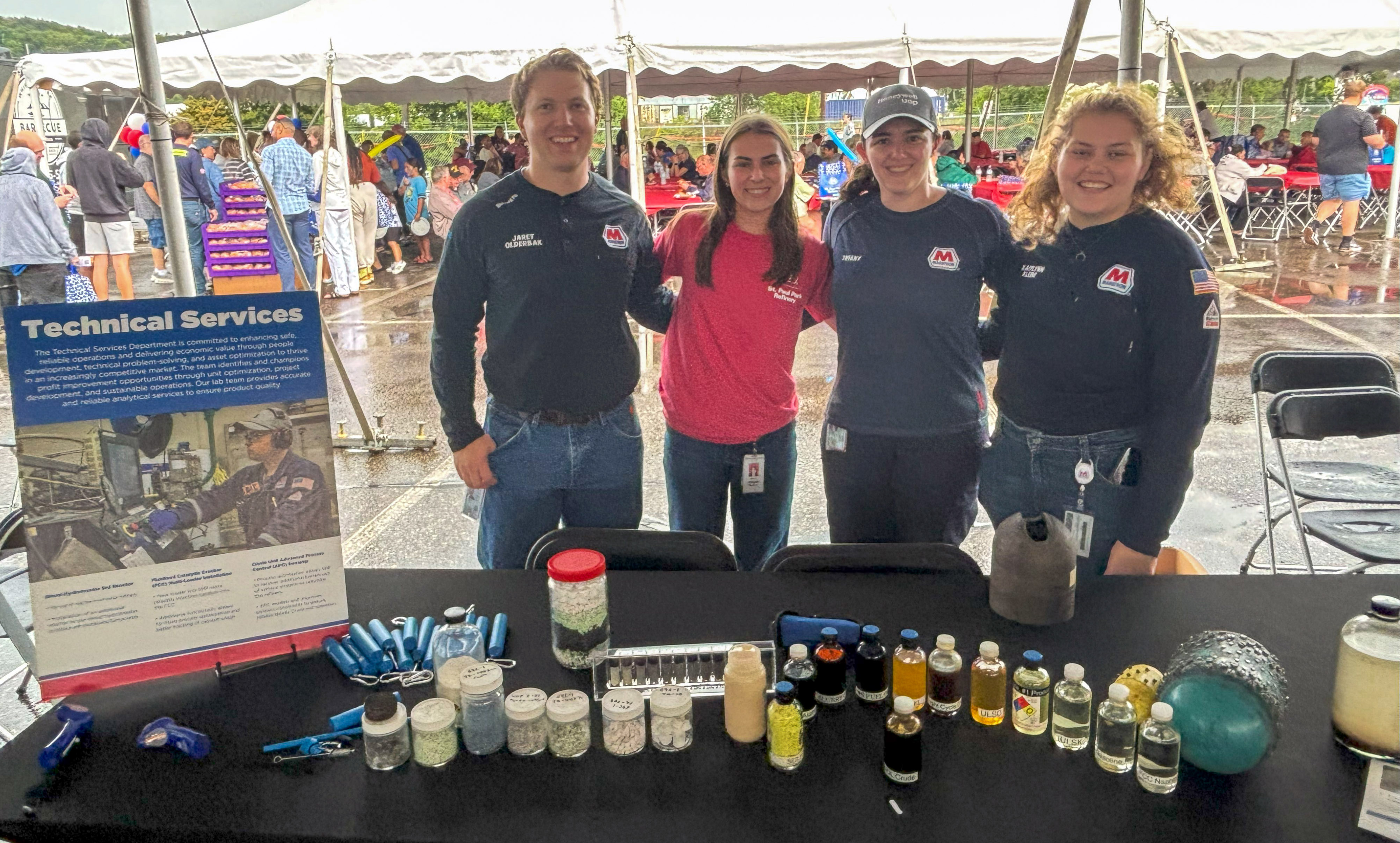 Four people stand behind a table under a tent, smiling for a photo.