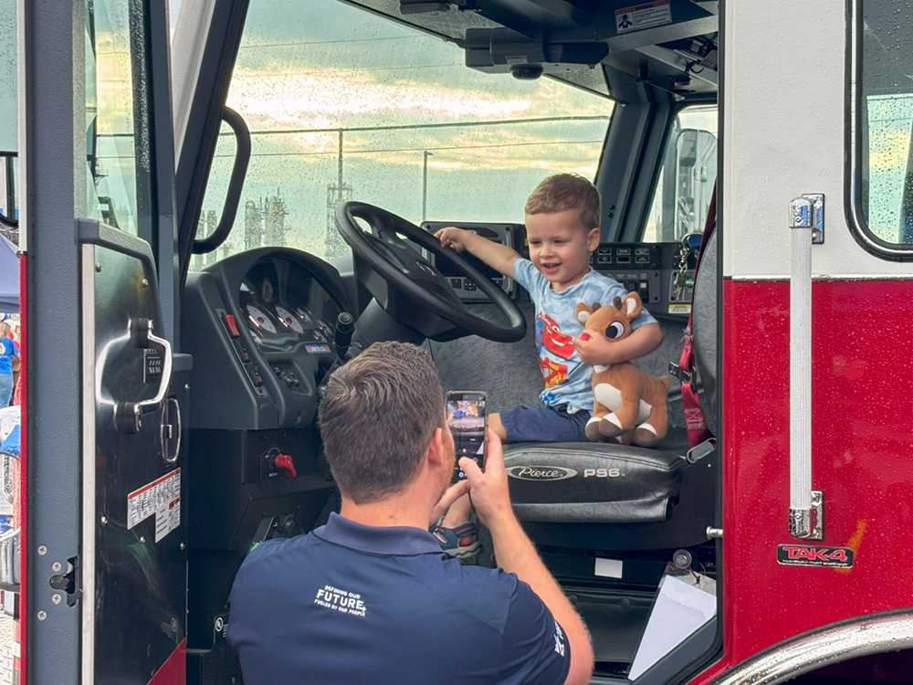 A young boy sits in the driver&rsquo;s seat of a fire truck while an adult takes his photo outside.
