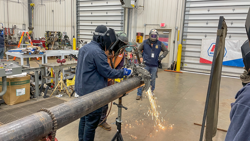 People in blue protective uniforms cut a pipe with a saw as sparks fly during a hands-on welding demonstration.