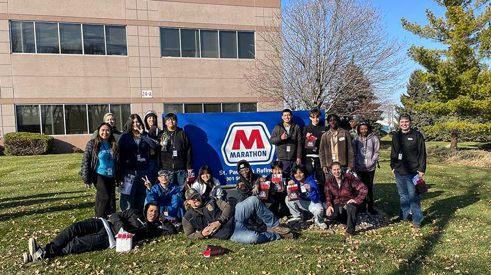 A group of people stand and lie on the grass outside next to a large sign.