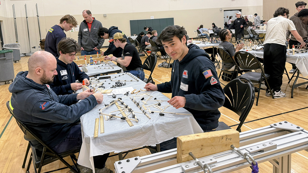 Volunteers and students sit at tables inside a gymnasium, working together on a hands-on project.