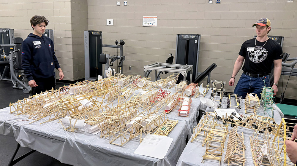 Two volunteers stand beside a display of student-built bridge designs inside a school gymnasium.