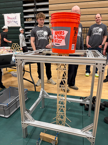 : A large orange bucket sits on top of a bridge structure inside a school gymnasium.